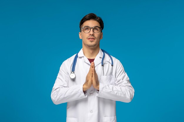 Journée des médecins beau mec mignon brune en blouse médicale priant et levant les yeux