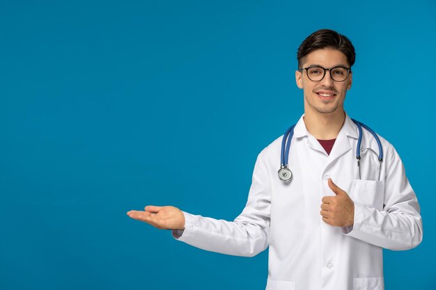 Journée des médecins beau mec mignon brune en blouse médicale souriant et montrant un bon geste