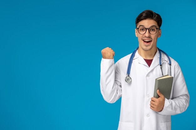 Journée des médecins beau mec mignon brune en robe médicale heureux tenant un livre