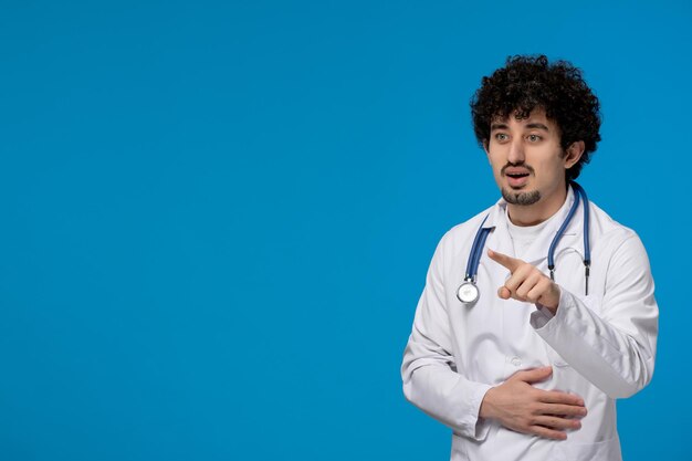 Journée des médecins bouclés beau mec mignon en uniforme médical pointant vers l'avant