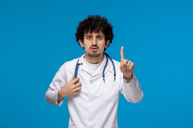 Journée des médecins bouclés beau mec mignon en uniforme médical regardant sérieusement et faisant arrêter le doigt
