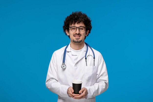 Journée des médecins bouclés beau mec mignon en uniforme médical souriant tenant une tasse en papier