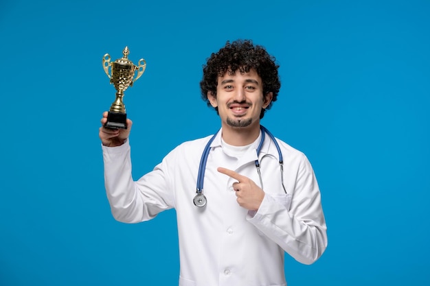 Journée des médecins bouclés beau mec mignon en uniforme médical souriant et tenant un trophée