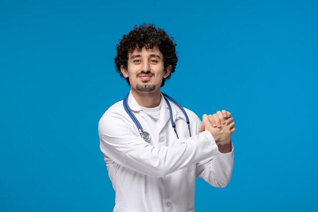 Journée des médecins bouclés beau mec mignon en uniforme médical tenant les mains ensemble et souriant