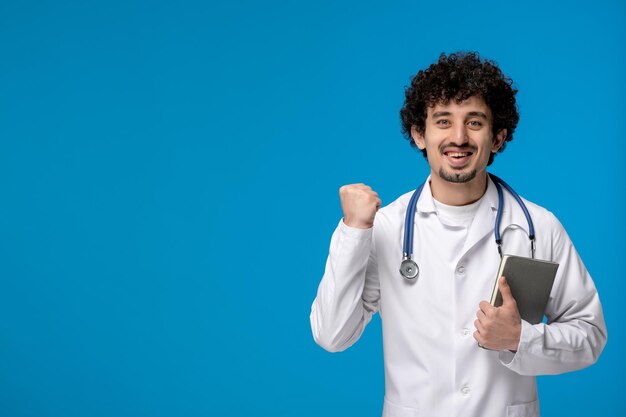 Journée des médecins bouclés beau mec mignon en uniforme médical tenant le poing levé et un livre