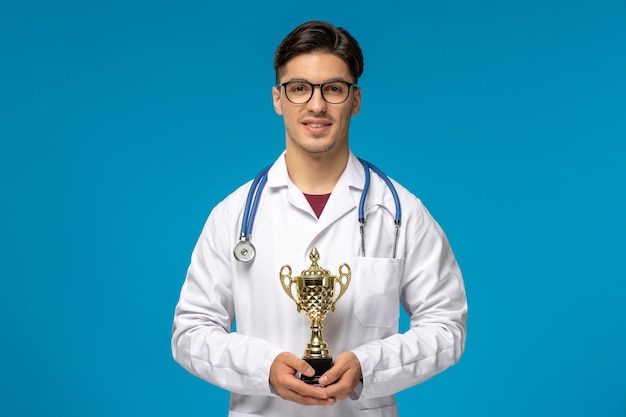 Journée des médecins mignon jeune bel homme en blouse de laboratoire et lunettes avec stéthoscope et trophée d'or