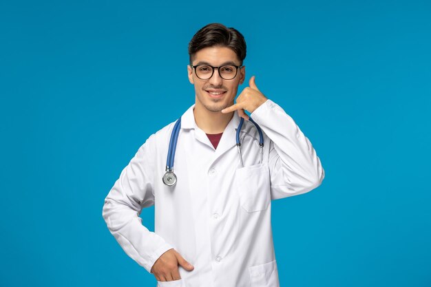 Journée des médecins mignon jeune mec brune en blouse de laboratoire portant des lunettes faisant un geste d'appel téléphonique