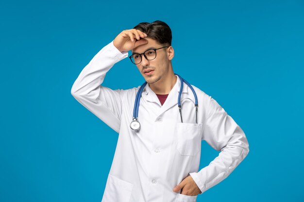 Journée des médecins mignon jeune mec brune en blouse de laboratoire portant des lunettes fatigué de toucher le front