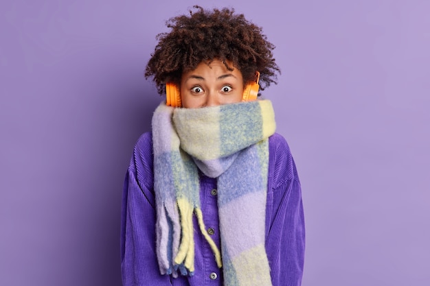 Photo gratuite joyeuse adolescente aux cheveux bouclés enveloppé dans une écharpe passe du temps libre à marcher en plein air pendant la journée d'hiver écoute une mélodie agréable.