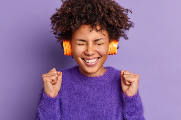 Joyeuse adolescente optimiste aux cheveux afro lève les poings serrés porte un pull décontracté écoute la musique préférée dans les écouteurs célèbre le jour de la chance exprime le bonheur