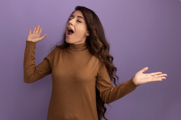 Joyeuse aux yeux fermés jeune belle fille portant un pull à col roulé marron répandant les mains isolées sur le mur violet