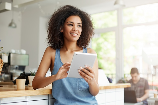 Joyeuse belle étudiante africaine au repos dans un café souriant à la recherche de côté tenant la tablette.