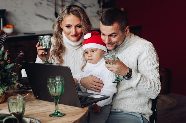 Joyeuse belle famille avec un bébé élevant des boissons tout en communiquant via un ordinateur portable depuis la maison.