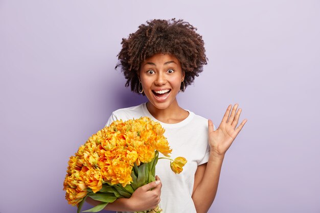 Joyeuse belle femme ravie tient des tulipes jaunes aromatiques, des vagues avec la paume, salue des amis, étant reconnaissant pour les félicitations, a une coupe de cheveux afro, porte un t-shirt blanc, des modèles sur un mur violet