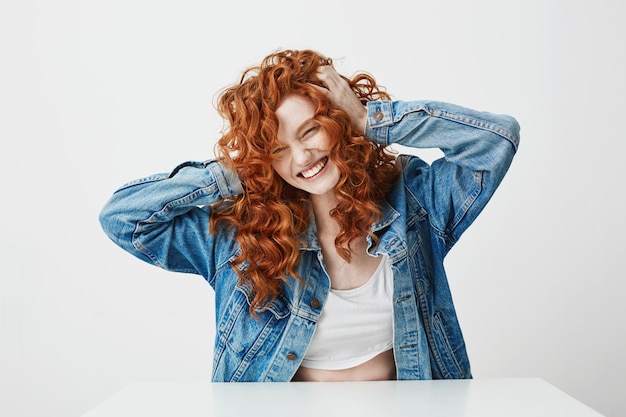 Joyeuse belle fille riant avec les yeux fermés touchant ses cheveux bouclés rouges.