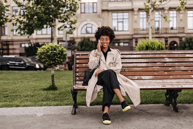 Joyeuse dame bouclée en pantalon noir et trench-coat beige est assise sur un banc en bois à l'extérieur