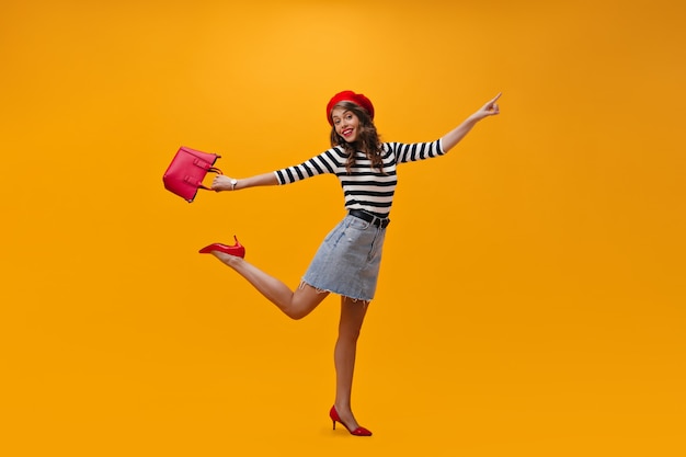 Joyeuse dame en chemise rayée, béret rouge sautant.n fond orange. avec sac à main. Jolie jeune femme en béret et chaussures à la mode posant.