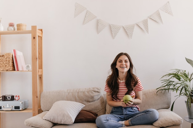 Joyeuse dame en jeans et t-shirt rit, s'assoit sur le canapé et tient la pomme