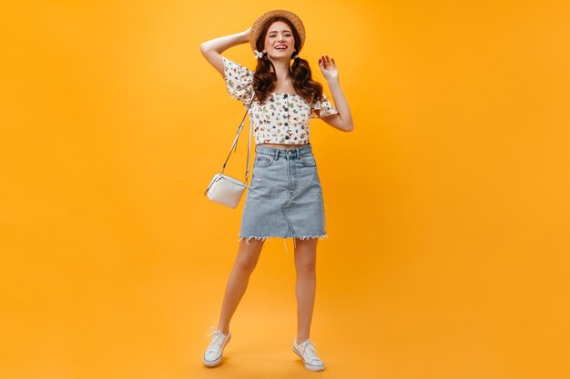 Joyeuse dame vêtue d'une jupe en jean et d'un haut court posant avec un sac blanc sur fond orange.