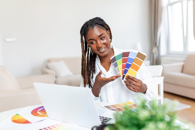 Joyeuse designer afro-américaine ayant une vidéoconférence avec des clients assis au bureau devant un ordinateur tenant des palettes de couleurs gesticulant et souriant copie espace