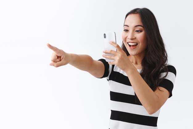 Joyeuse divertie enthousiaste jeune femme asiatique joyeuse en tenue décontractée enregistrer une vidéo en vérifiant quelque chose d'incroyable pointant le doigt sur le côté en photographiant un concert éclairé sur fond blanc