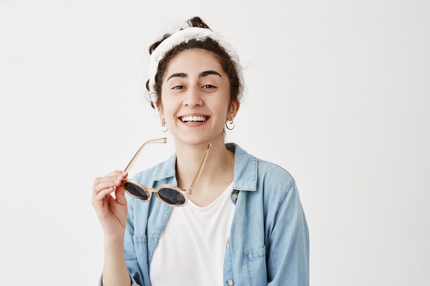 Joyeuse étudiante aux cheveux noirs en do-rag sourit largement, montre des dents blanches et uniformes, vêtue d'une chemise en jean, passe une excellente journée, se détend à l'intérieur isolé contre un mur blanc avec un espace de copie.