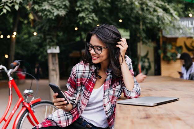 Joyeuse étudiante jouant avec ses cheveux noirs. Portrait en plein air de fille blithesome tenant le téléphone alors qu'il était assis sur la rue.