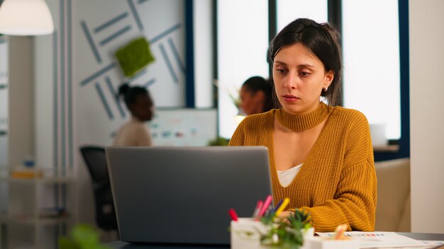 Joyeuse femme d'affaires tapant sur un ordinateur portable et souriante assise au bureau dans un bureau de démarrage occupé profitant du travail dans un lieu de travail créatif. Une équipe diversifiée analyse les données statistiques dans une entreprise moderne