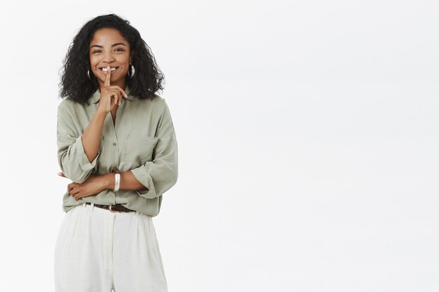 Joyeuse femme afro-américaine charismatique et élégante avec une coiffure frisée shushing et souriant avec un regard joyeux disant secret