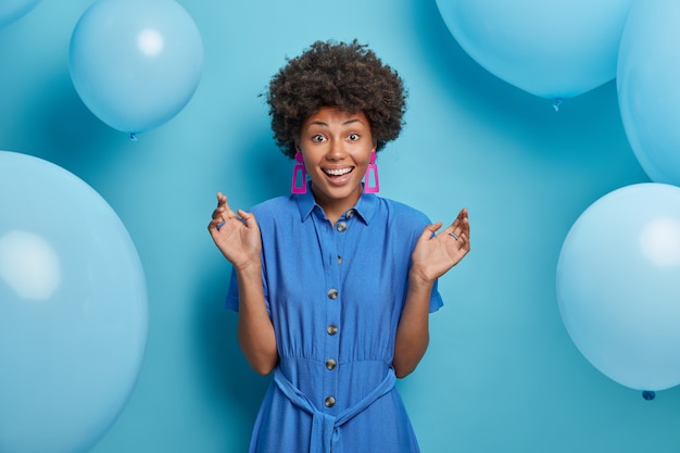 Photo gratuite joyeuse femme afro-américaine heureuse de rencontrer des amis à la fête, se tient avec les mains levées sourit largement, célèbre son anniversaire, porte une robe bleue, a hâte d'ouvrir des cadeaux, a de nombreux ballons à air autour