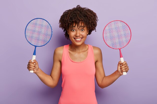 Joyeuse femme afro-américaine tient deux raquettes de tennis, invite à la rejoindre et jouer au jeu, se repose entre les matchs de tennis