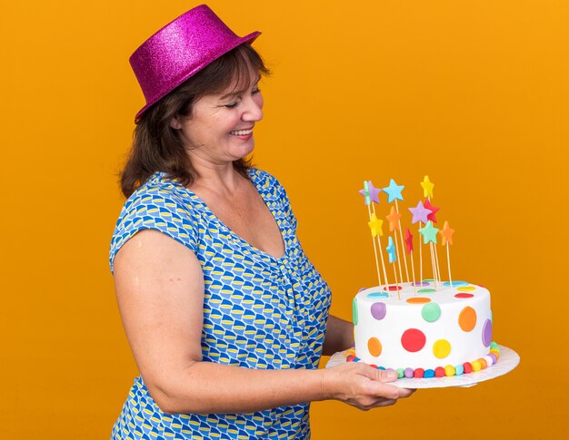 Joyeuse femme d'âge moyen en chapeau de fête tenant un gâteau d'anniversaire en le regardant avec le sourire sur le visage