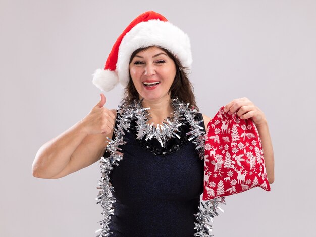 Joyeuse femme d'âge moyen portant bonnet de Noel et guirlande de guirlandes autour du cou tenant le sac de cadeau de Noël regardant la caméra montrant le pouce vers le haut isolé sur fond blanc