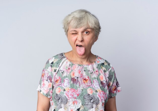 Joyeuse femme âgée clignote des yeux et sort la langue isolée sur un mur blanc