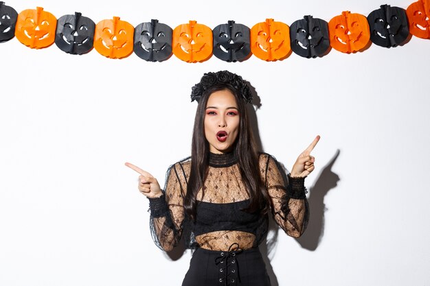 Joyeuse femme asiatique en robe de dentelle gothique célébrant l'halloween, pointant du doigt sur le côté, debout sur fond blanc avec des banderoles de fête de citrouille.