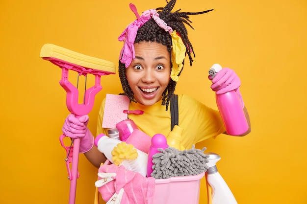 Photo gratuite joyeuse femme au foyer ethnique a des poses de tresses avec une vadrouille et une bouteille de détergent heureuse de nettoyer