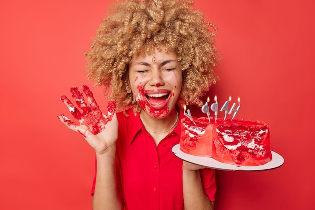 Joyeuse femme aux cheveux bouclés folle autour de sale avec de la crème mange un délicieux gâteau sucré avec les mains garde la paume levée s'exclame de joie porte une robe isolée sur fond rouge vif Dessert manger