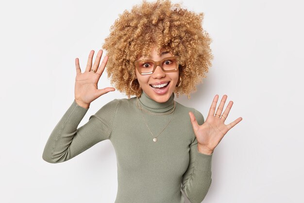 Joyeuse femme aux cheveux bouclés ludique lève les paumes se sent heureux sourit largement porte des lunettes col roulé décontracté essaie d'amuser quelqu'un isolé sur fond de studio blanc. Concept d'émotions heureuses