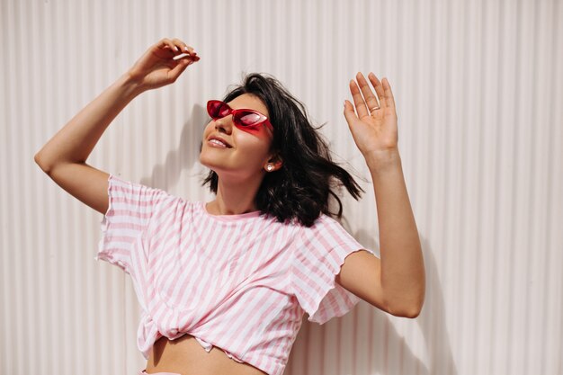 Joyeuse femme aux cheveux courts bénéficiant d'une journée ensoleillée. Modèle féminin détendu dans des lunettes de soleil debout avec les mains sur fond texturé.