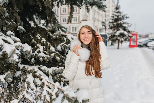 Joyeuse femme blanche porte un bonnet tricoté et un manteau chaud posant avec un sourire doux à côté de l'arbre. Modèle féminin extatique aux cheveux longs en veste avec fourrure profitant des vacances d'hiver en plein air.
