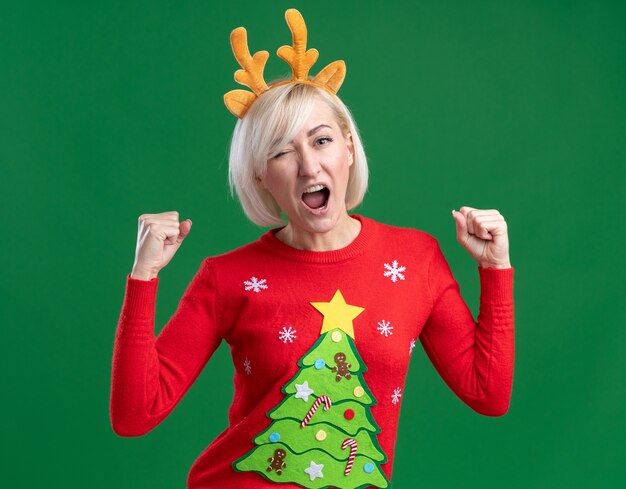 Joyeuse femme blonde d'âge moyen portant bandeau de bois de renne de Noël et chandail de Noël faisant un clin d'œil faisant oui geste isolé sur mur vert