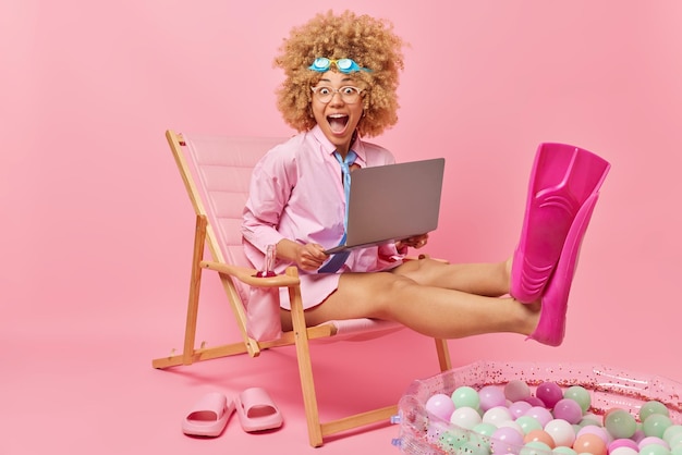 Photo gratuite joyeuse femme bouclée surprise rêve de vacances d'été porte une chemise à lunettes et des palmes fonctionne sur un ordinateur portable pose sur une chaise longue sur fond rose concept de tourisme de vacances de personnes