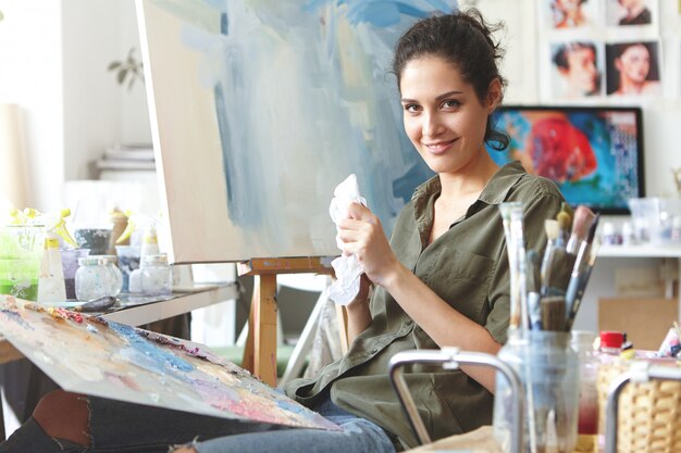 Joyeuse femme brune assise à l'atelier, dessin d'image colorée sur chevalet, à l'aide d'huiles brillantes. Peintre professionnel étant heureux d'avoir l'inspiration pour la peinture. Profession, concept d'occupation