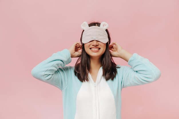 Joyeuse femme brune en pyjama doux blanc et menthe met un joli masque de sommeil et sourit largement sur fond rose isolé