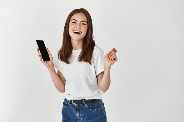 Joyeuse femme brune riant et utilisant un téléphone portable, debout heureuse avec un téléphone portable sur un mur blanc