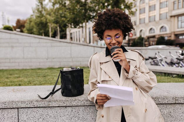 Joyeuse femme brune en trench-coat beige élégant boit du café à l'extérieur