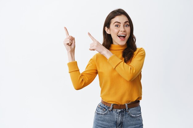 Joyeuse femme candide pointant vers le logo du coin supérieur gauche et souriante, montrant une bannière promotionnelle sur le dessus, debout contre un mur blanc