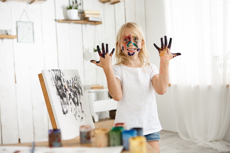 Photo gratuit joyeuse femme caucasienne kid démontrant ses mains en peinture noire, debout derrière le chevalet avec sa photo.