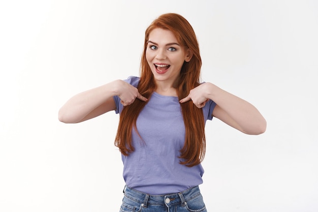 Joyeuse femme caucasienne rousse amusée et souriante en t-shirt violet, pointant sa poitrine et souriant, parlant de réalisation personnelle, se vantant, faisant du bénévolat, suggérant une candidature, mur blanc