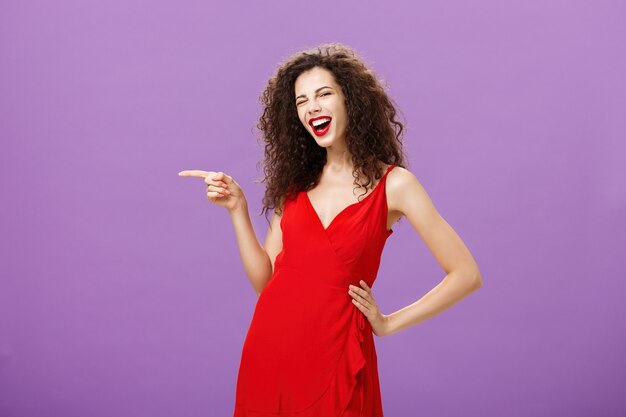 Joyeuse femme élégante et insouciante avec une coiffure frisée s'amusant lors d'une fête géniale dans un dre luxueux rouge ...
